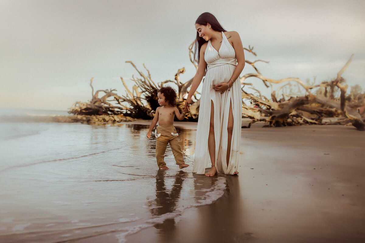 A woman in a flowing white dress stands on a beach, her hand gently clasping that of a young child. As driftwood frames the scene and gentle waves kiss their feet, the moment captures the essence of a Macon beach maternity photographer's dream.