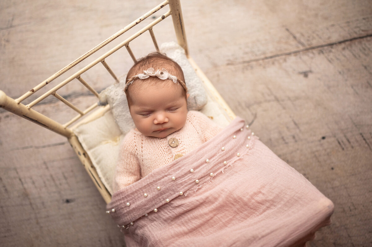 Portrait of newborn girl asleep on tiny doll bed covered with pink, pearl embellished blanket.