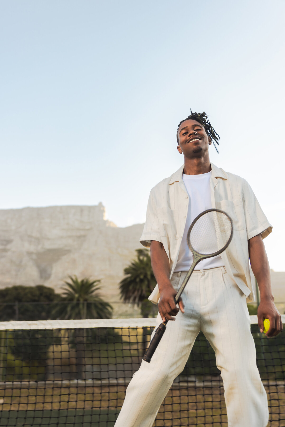 Man in a cream-colored outfit smiling confidently on a tennis court, holding a tennis racket and ball, with Table Mountain and palm trees in the background.