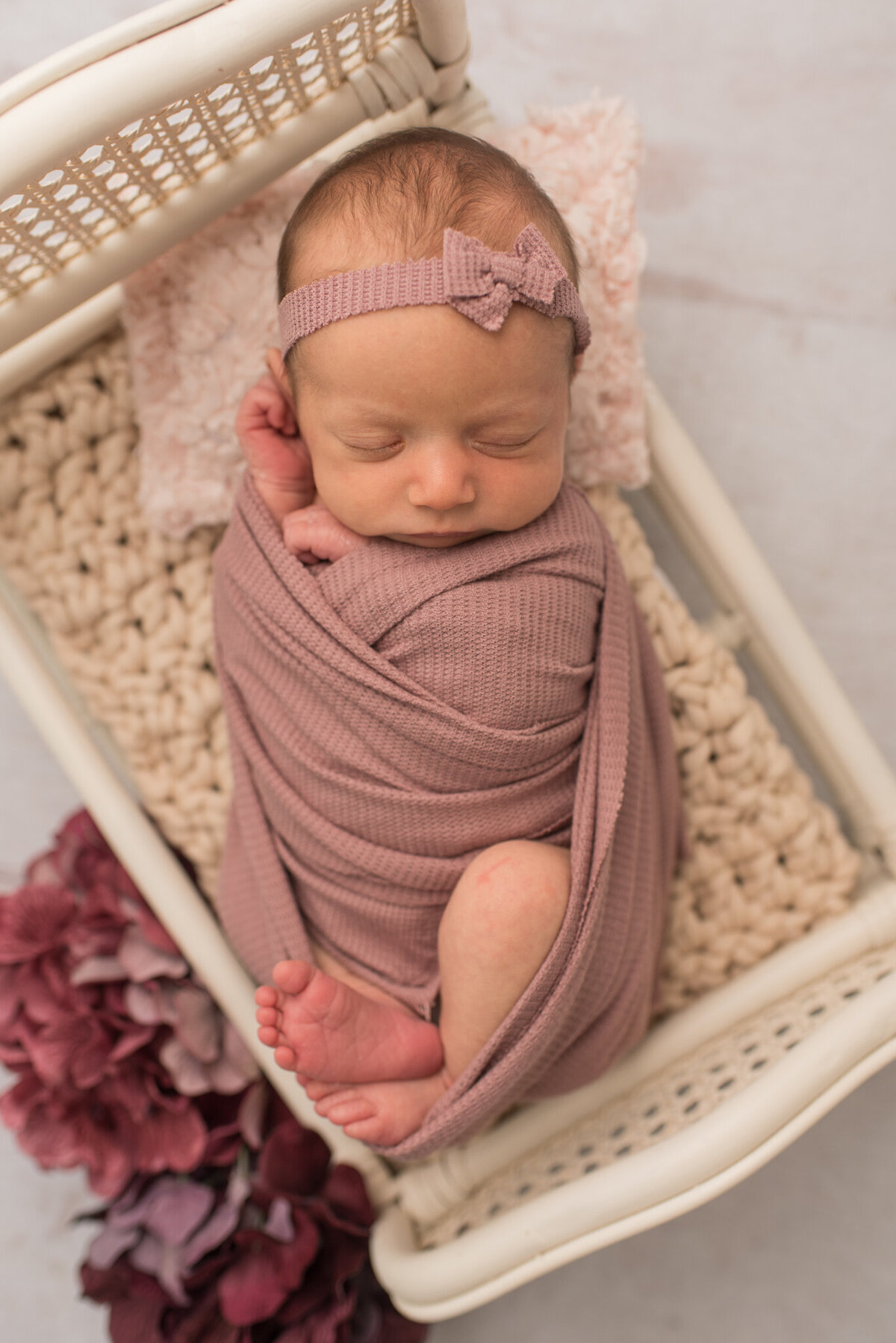 A newborn baby girl is wrapped in a mauve wrap with her head on a pink pillow with bow in her hair.
