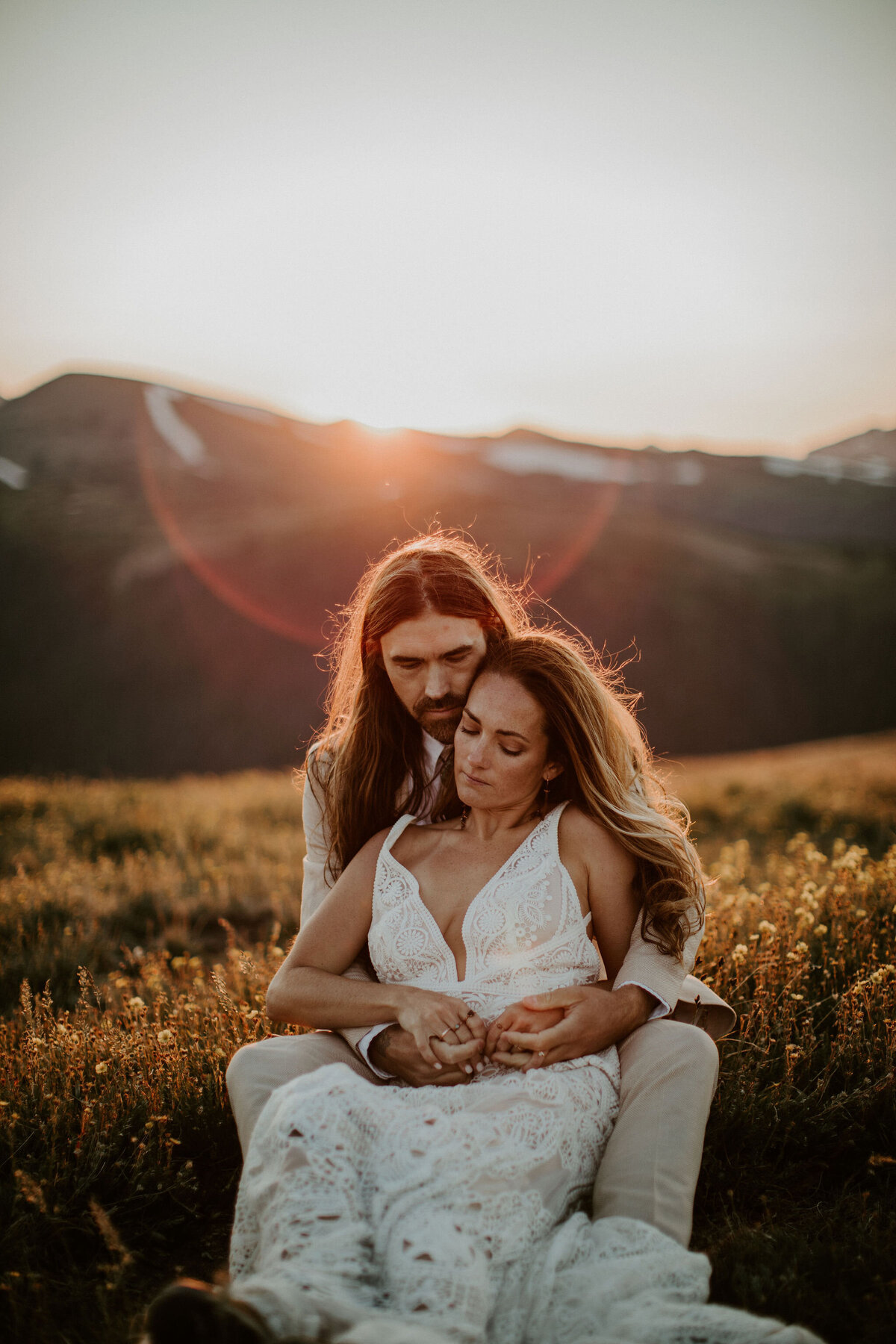 Bride and groom wearing an ivory suit and white wedding gown sit in a field holding hands during golden hour.
