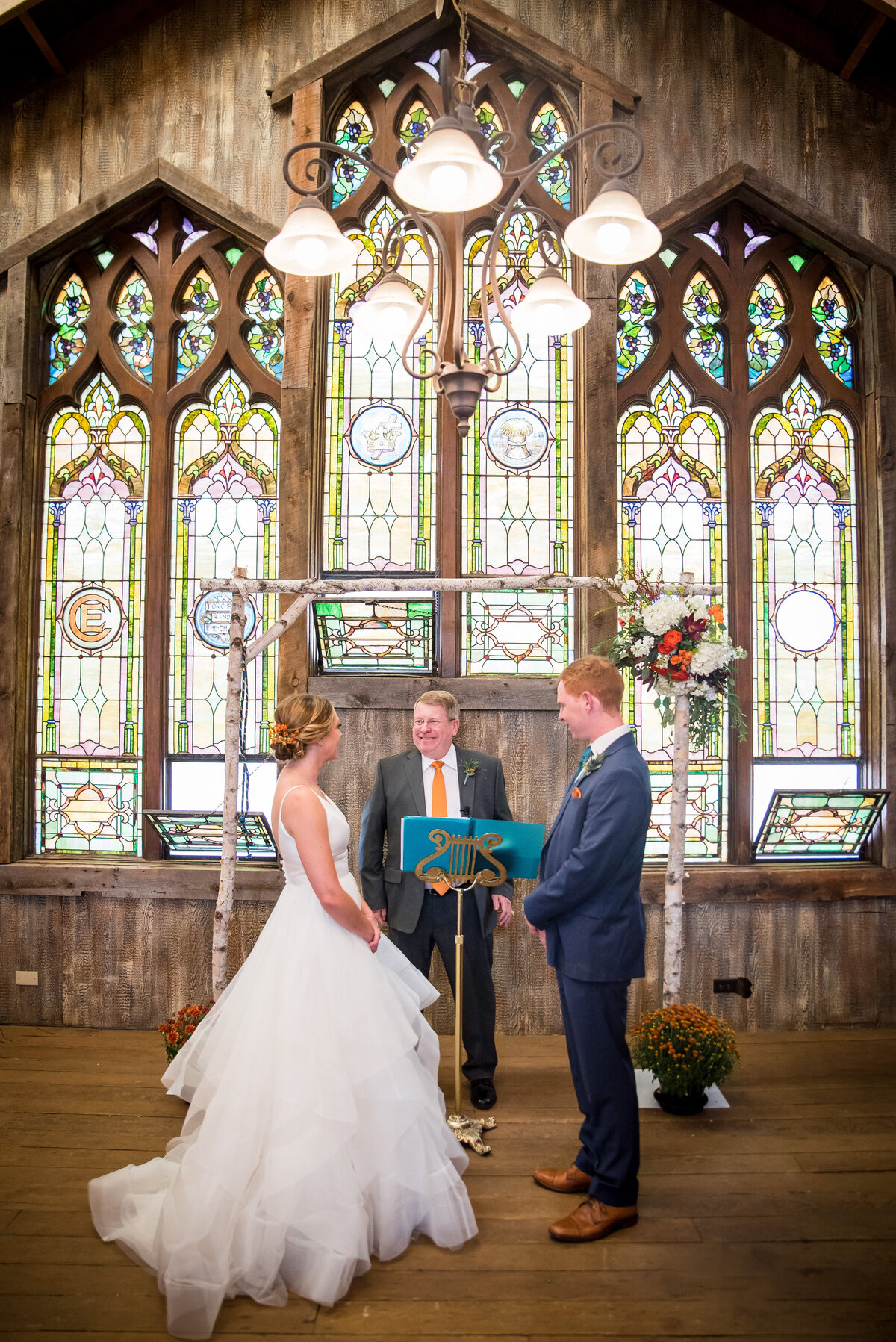 A bride, groom, and officiant stand at the front of a chapel during the ceremony with ornate stained glass windows in the background.