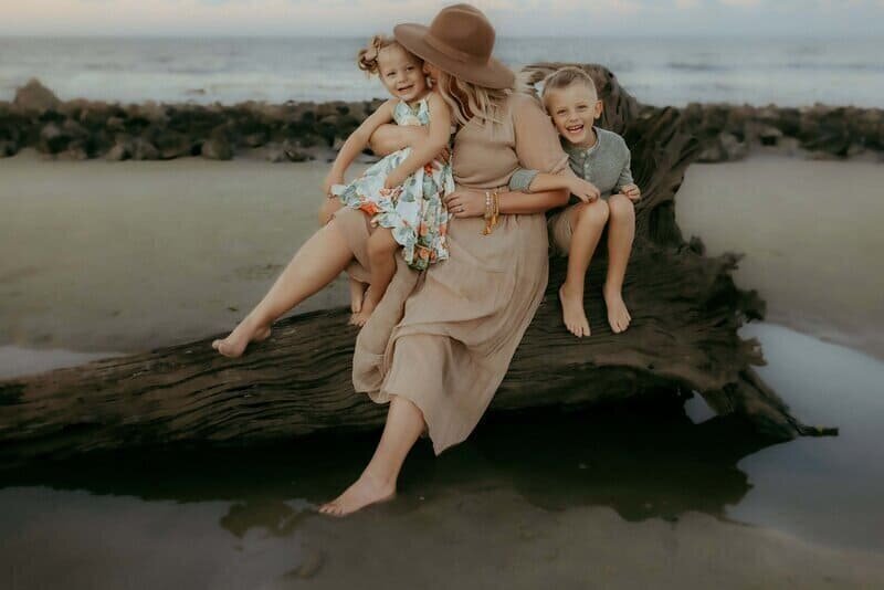 A woman wearing a hat sits on a driftwood log with two smiling children at the beach.