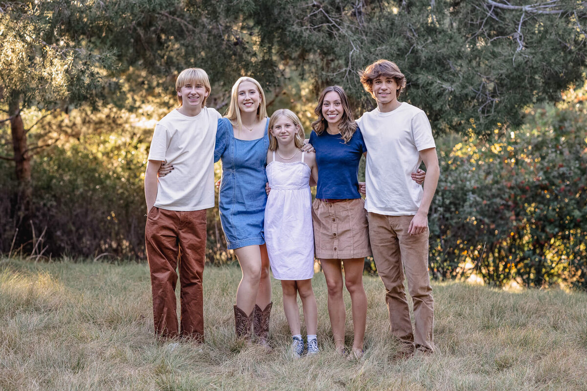 Five young adult cousins with blonde and brunette hair, smiling with their arms around each other in a park.
