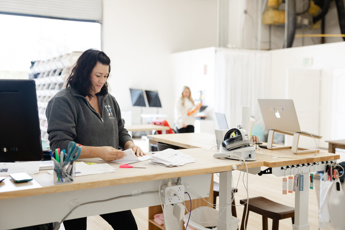 Woman with dark hair working on embroidery machines in bright studio. Photograph by Yucaipa branding photographer Kaitlyn Dawn Photography.