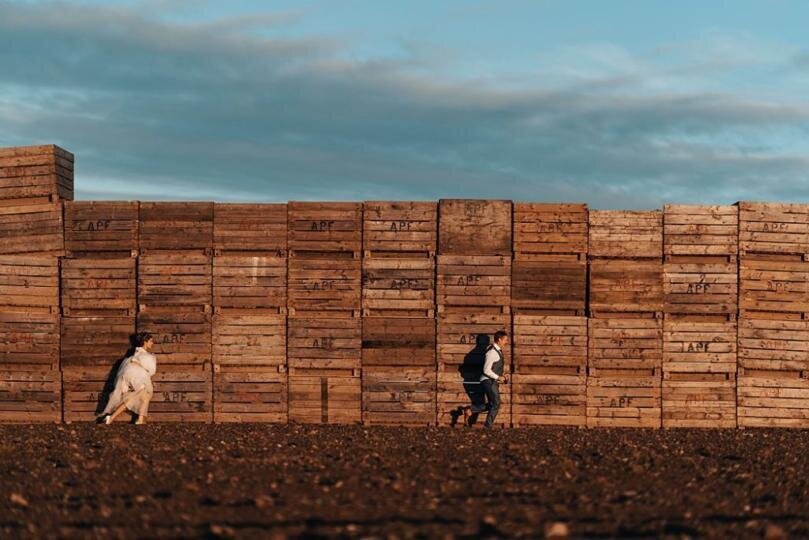 Bride in bridal gown chasing the Groom. Both running in to the right in front of a wall of wooden crates in a field