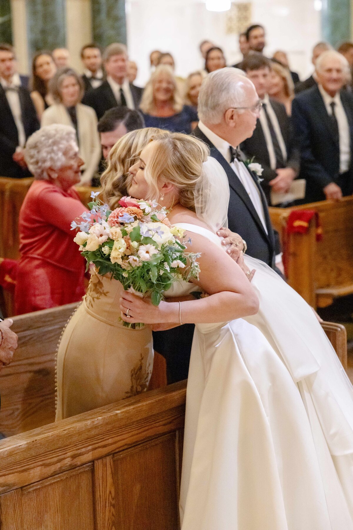 A bride in a white dress hugs a woman holding a bouquet of flowers in a church, surrounded by standing guests. The scene captures an emotional moment during a wedding ceremony.