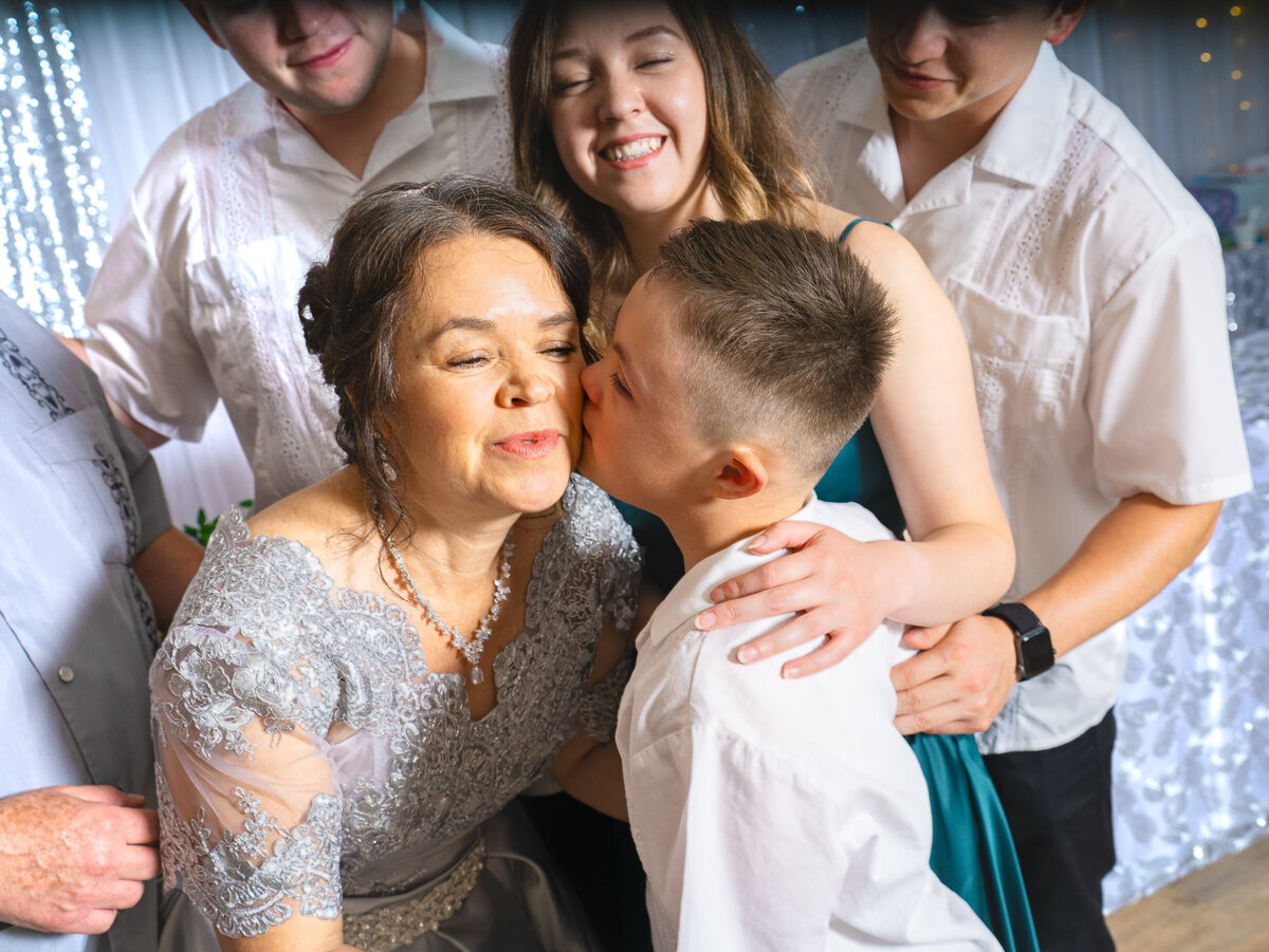 A mature bride in a silver wedding dress, surrounded by her children in a sparkling ballroom, leans down to receive a kiss on the cheek from her young son.  Photo by SAVI Photography - Los Angeles CA Photographer