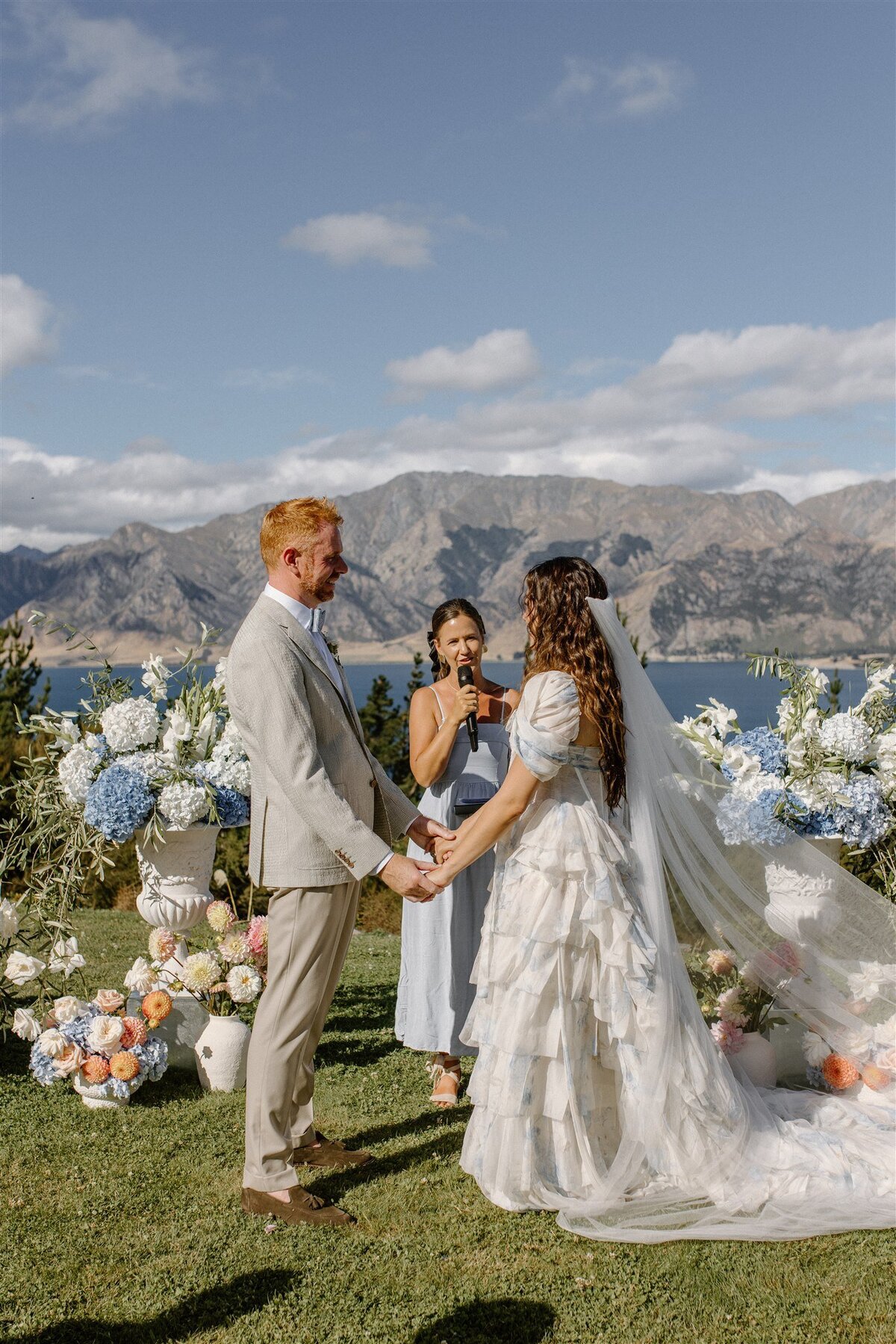 Wedding couple holding hands during their outdoor ceremony at Lake Hāwea, framed by blue and white florals and a celebrant in soft blue.