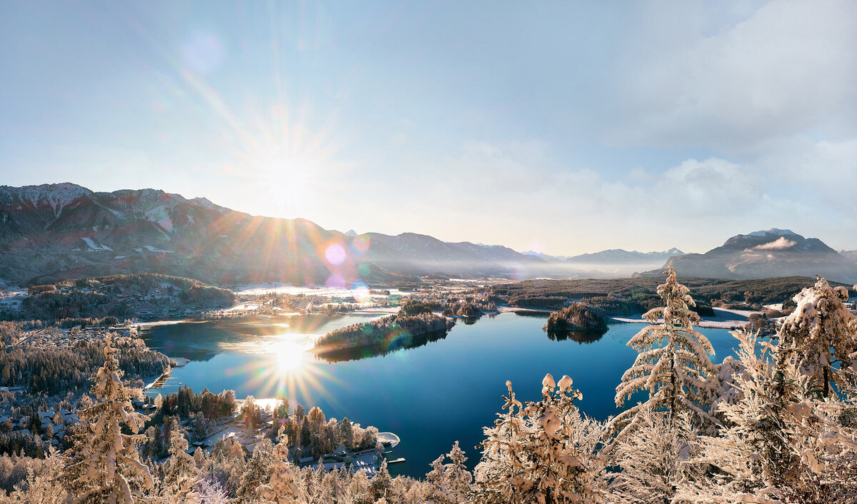Verschneite Aussicht auf den Faaker See im Morgenlicht mit Sonnenstern über den Bergen – winterliche Landschaftsaufnahme mit klarem Himmel und Lichtreflexen