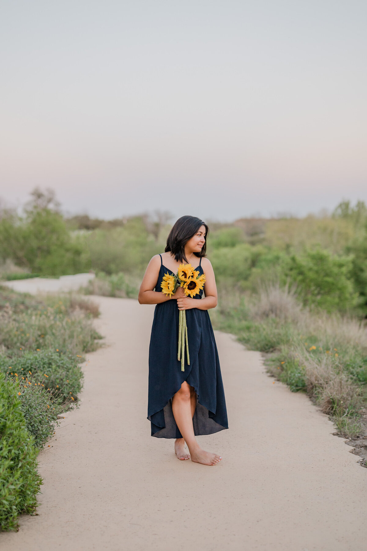 Girl in bare feet and a blue dress walks along  a path holding sunflowers during senior pictures by Cassey Golden Photography.