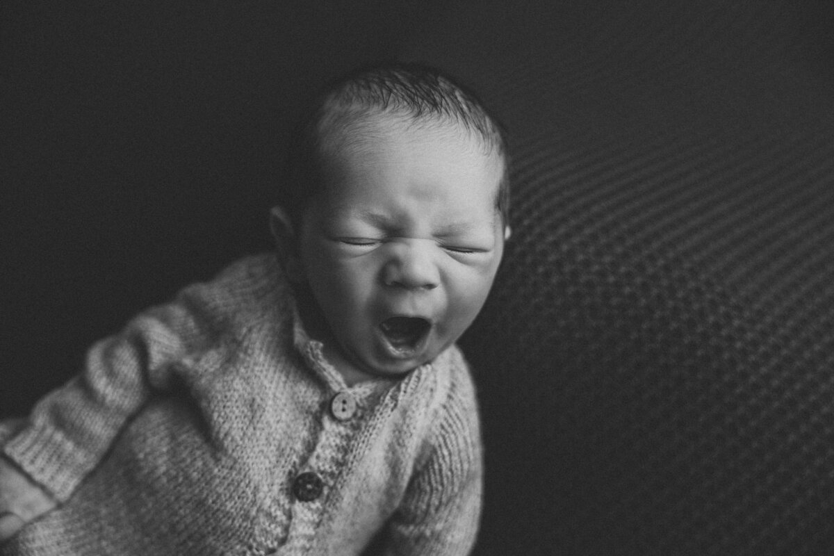 A black and white image of a newborn laying down yawning while wearing a sleeper.