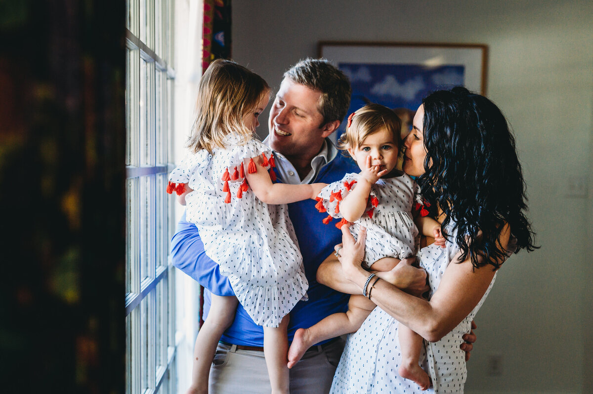 parents stand near window and snuggle young daughters at home