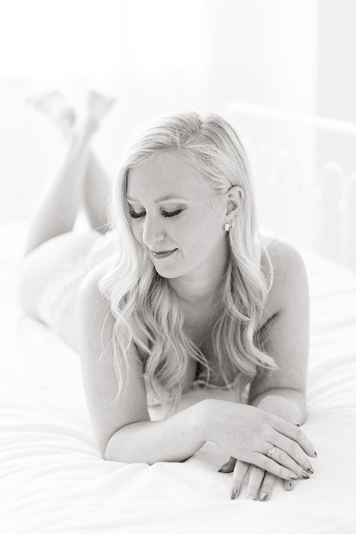 Black and white photo of a woman laying on a bed dressed in a matching white lace lingerie set as she poses for her boudour session while looking to the side of her shoulder.