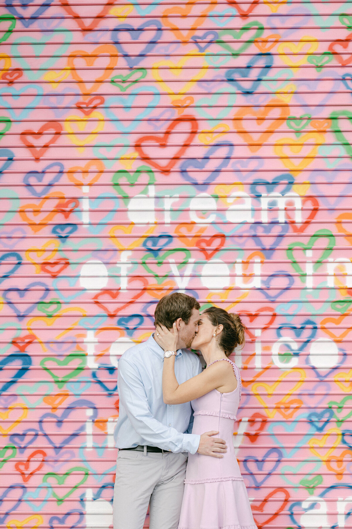 Engaged couple sharing a kiss in front of a colorful heart mural with 'I dream of you' text in Miami, captured by Rodrigo Varela Photography.