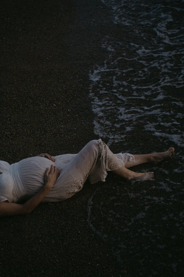 La future maman allongée sur le sable dans une robe blanche légère mouillée par les vagues. Séance intime par Kamescop.
