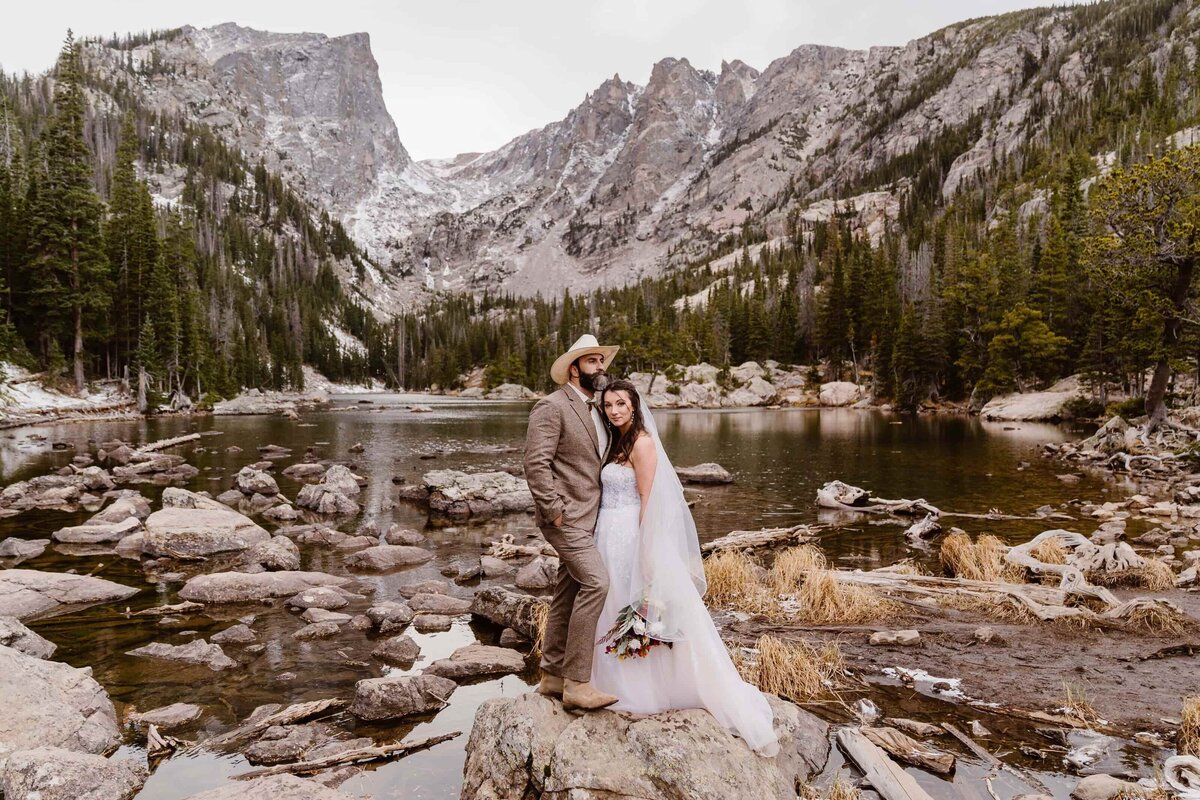Bride and groom standing on a rock by the water during their wedding at Dream Lake in Rocky Mountain National Park, with dramatic mountain peaks and evergreen trees in the background.
