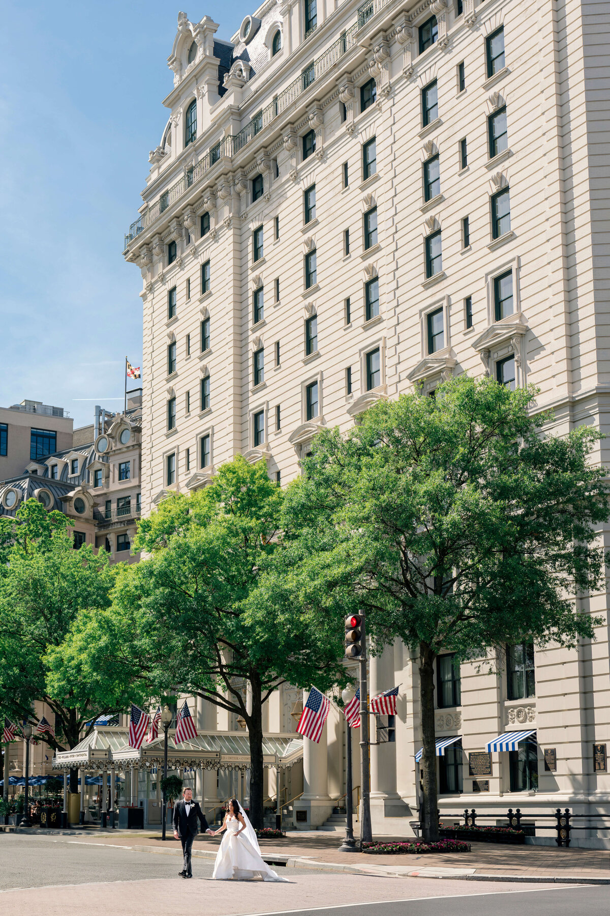 A bride and groom walk hand in hand across a street in front of a large, historic building with multiple floors and white stone facade. Several American flags are displayed. Green trees line the sidewalk under a clear, sunny sky.