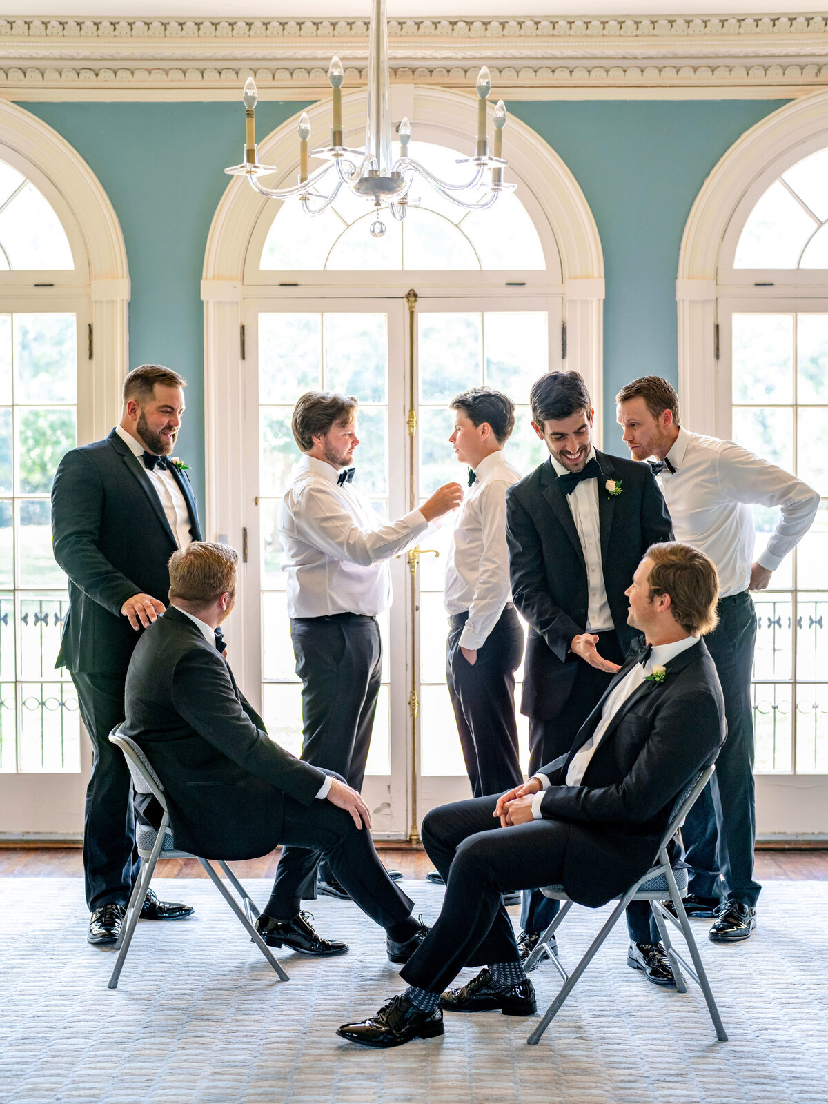 A group of seven men dressed in formal suits and bow ties are gathered in a room with large arched windows. Three men are seated on chairs while four are standing, engaging in conversation and adjusting each others attire. The room is elegant with muted blue walls.