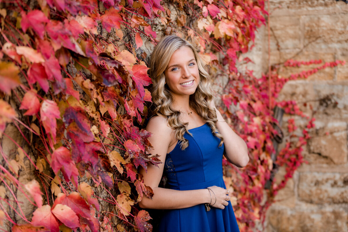 A girl in a blue dress leans against a red and orange ivy wall and smiles at the camera for her senior portrait.