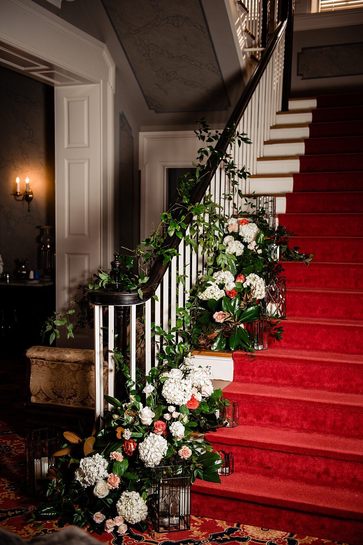 Red carpeted staircase at Rippa Villa decorated with a lush floral installation of white hydrangea, orange roses, peach roses, pink carnations and greenery climbing up the length of the white banister with a dark wood top at Rippa Villa mansion.