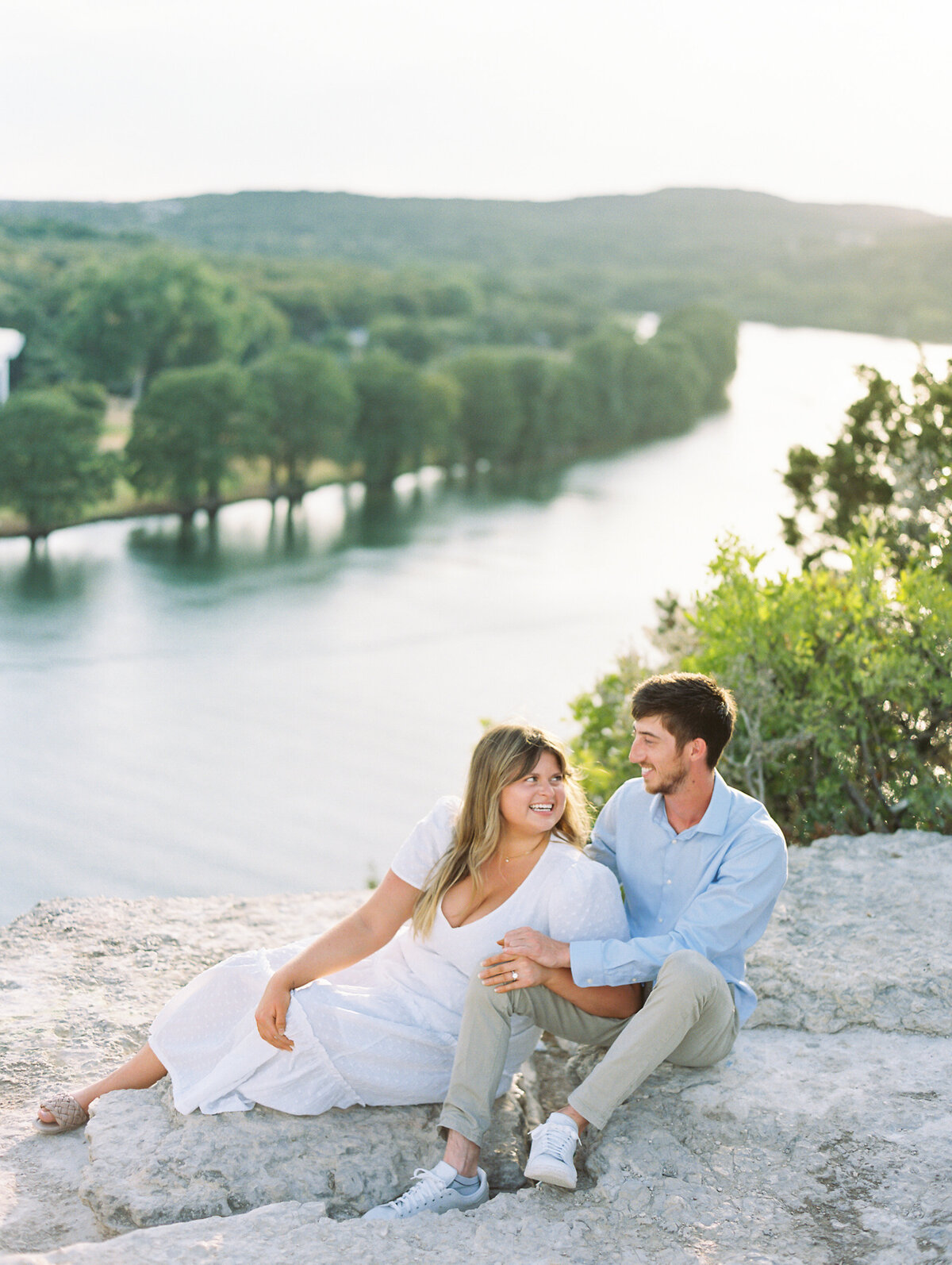 Woman in a white dress and man in a blue shirt posing on a rock in front of a landscape of water and trees