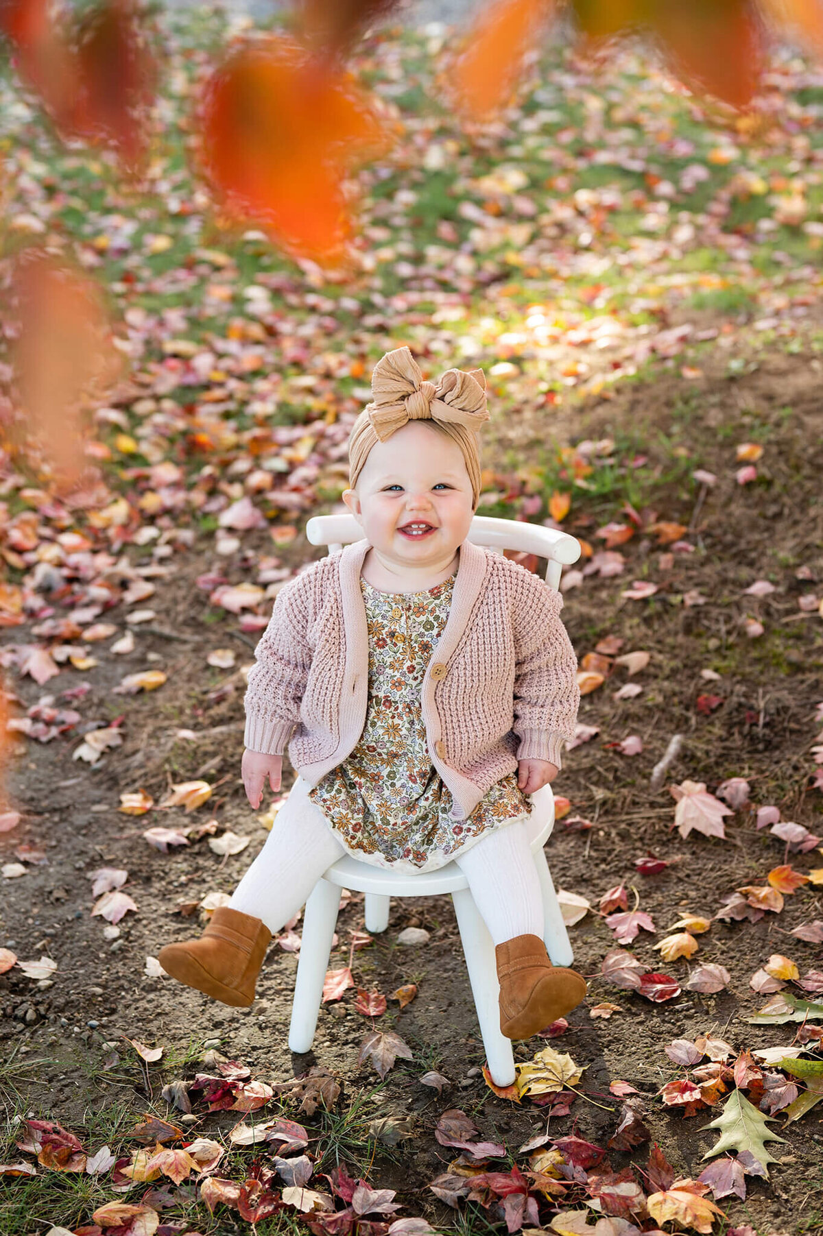 Baby sits on a white chair smiling at the camera with fall foliage around the edge of the photo.