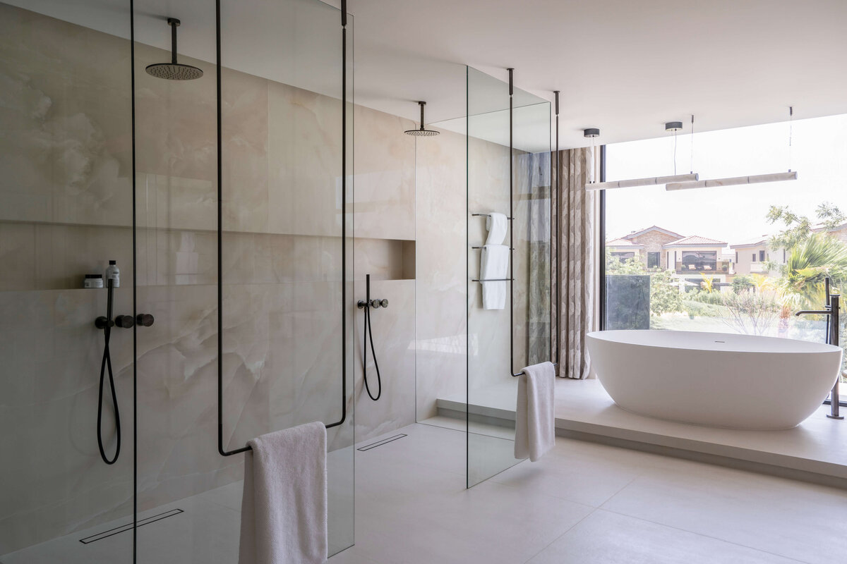 Large walk-in glass double shower with neutral marble walls and black hardware. To the right of the showers, a large soaking tub and window wall can be seen.