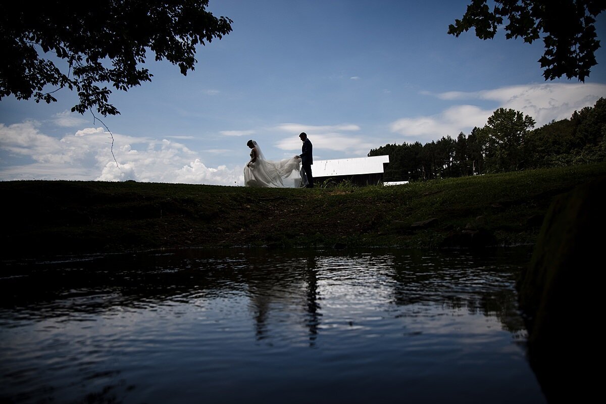 Bride wearing a long lace dress and veil being followed by the groom who is carrying the train of her dress as they walk along a hill at sunset with their images reflected in the pond below