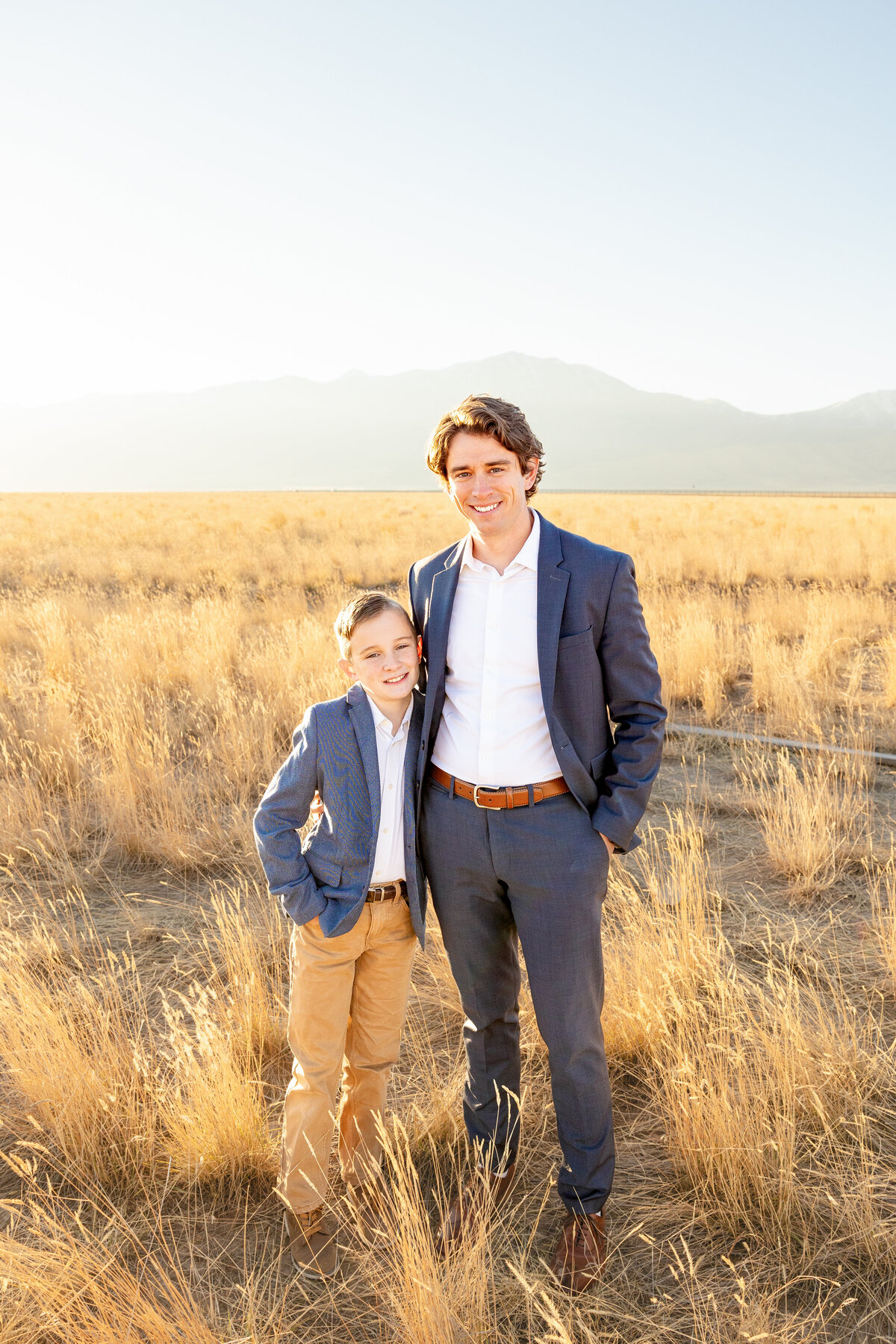 A father and son dressed in blue suits stand together in a golden field, smiling at the camera with mountains behind them.