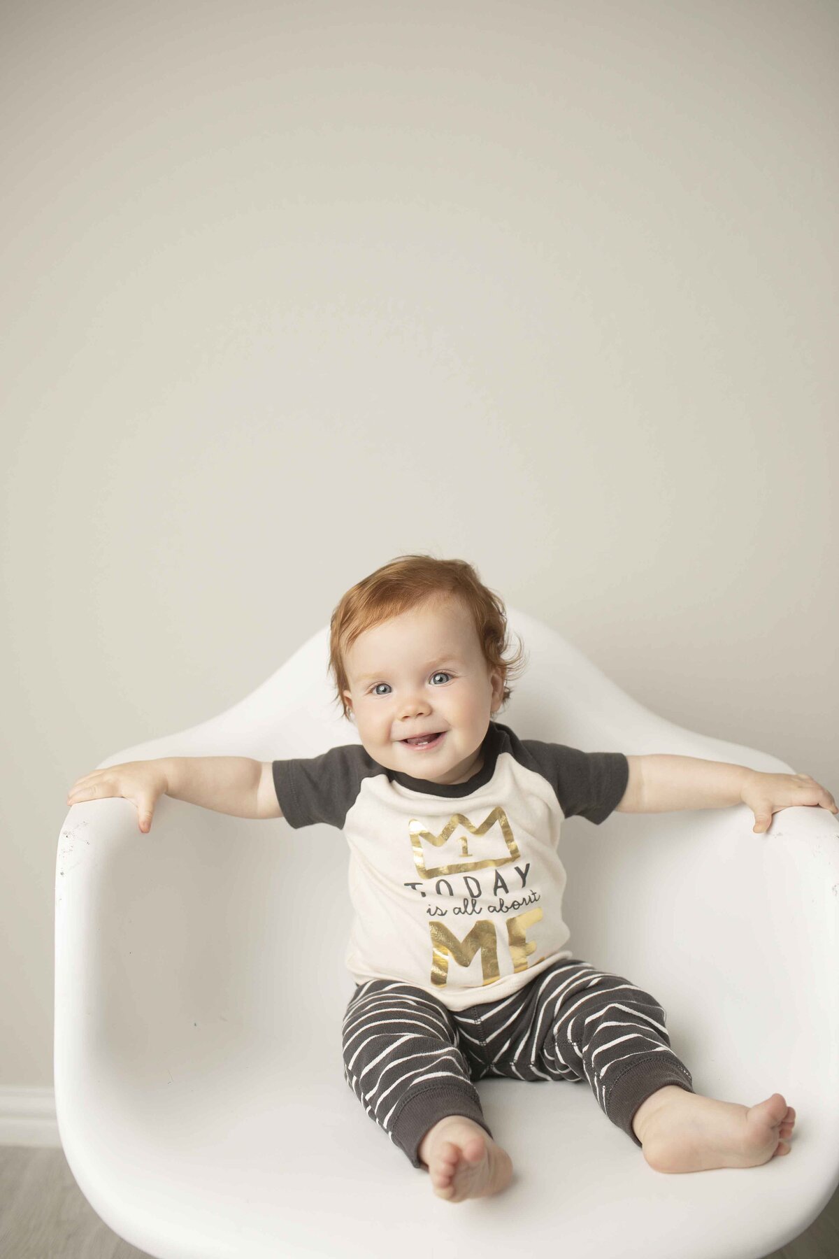 One-year-old redhead birthday boy in a white chair, looking playful and happy in a studio setting