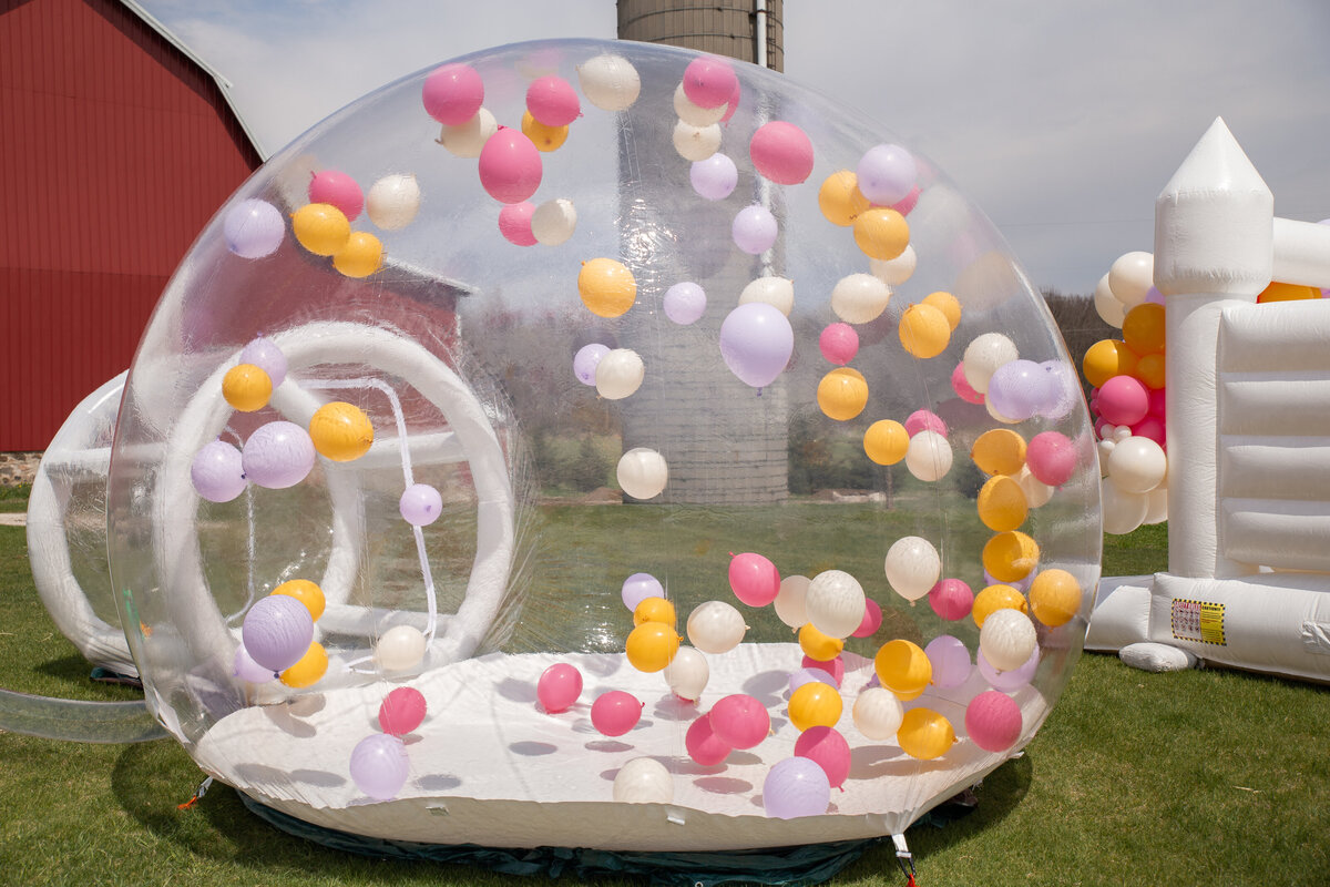 A large, clear bubble house set up outdoors with colorful balloons flying around inside.