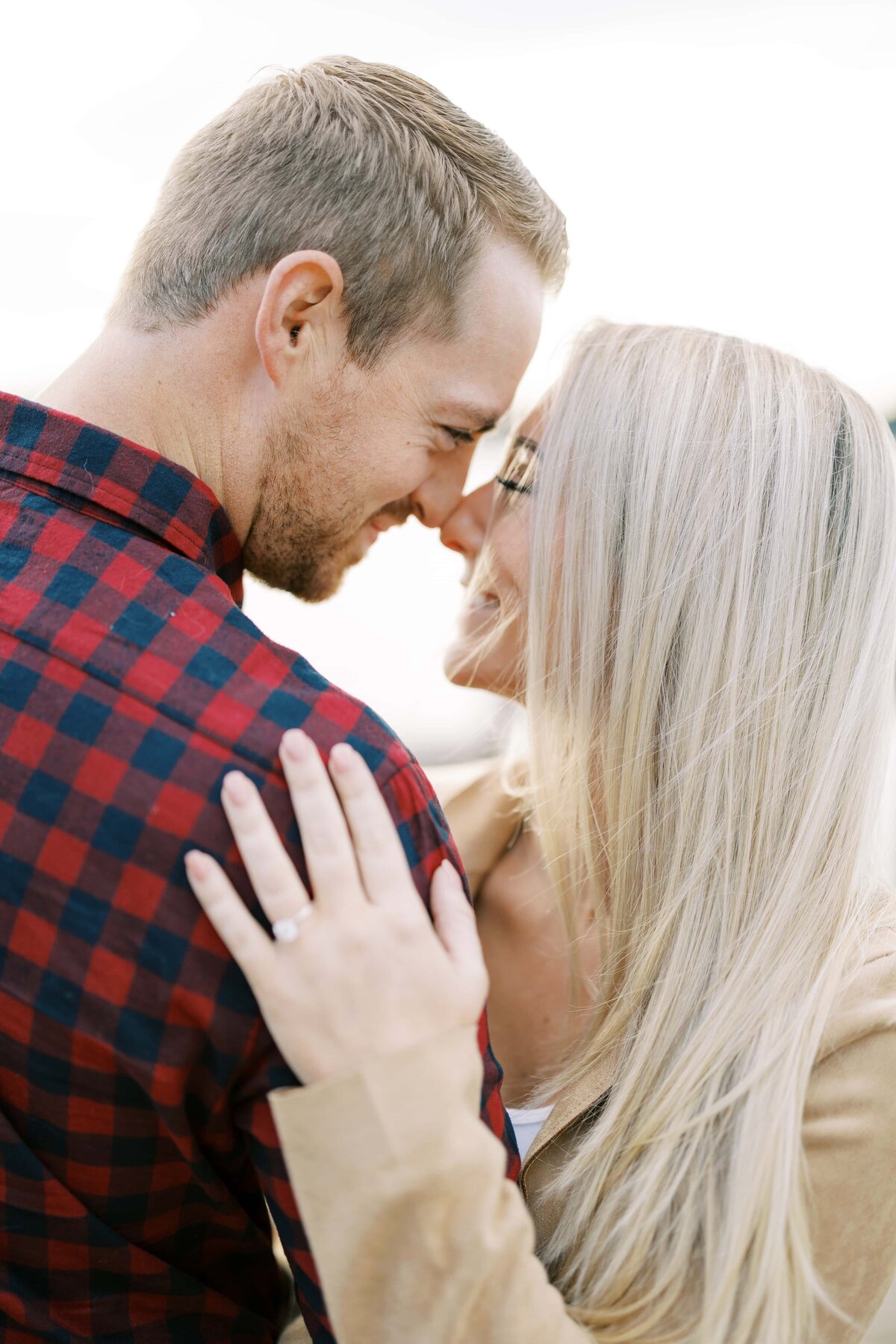 Man with buffalo plaid shirt smiling at woman with white blonde hair with their foreheads touching as she rests her hand on his shoulder featuring a large engagement ring.