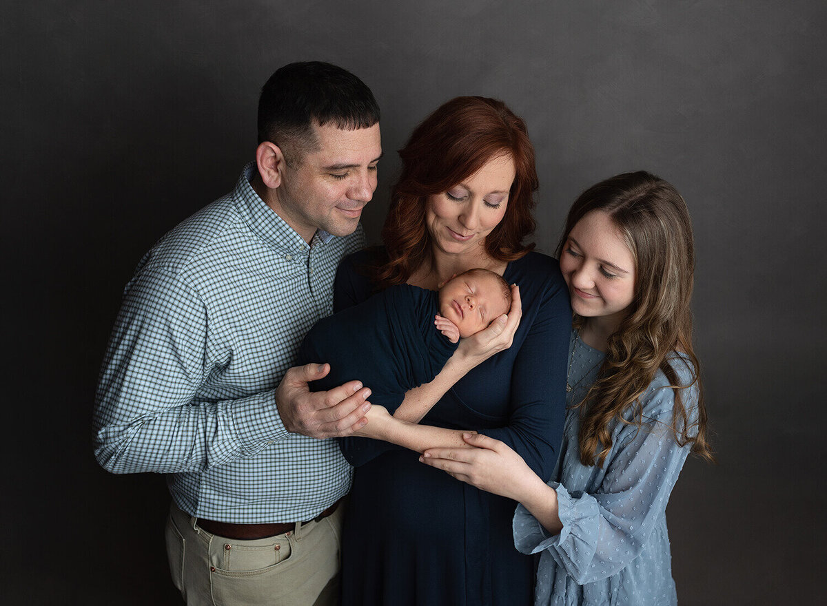Happy parents and their teen daughter cradle a sleeping newborn while standing in a studio