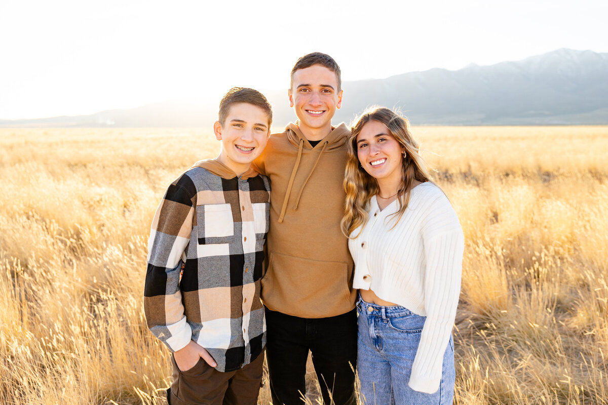 Three siblings stand together in a golden field in Eagle Mountain, Utah, smiling at the camera with the setting sun casting a warm glow on them.