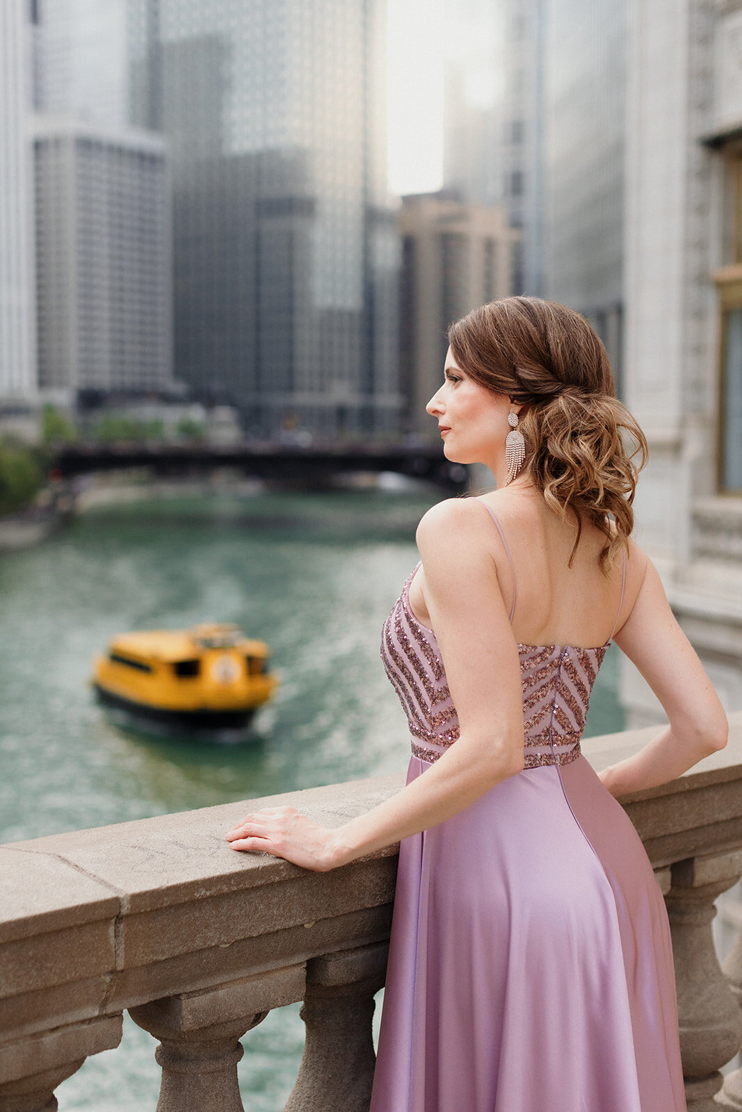 fair skinned woman in lavender gown is looking out over the Chicago River. A yellow boat is in the distance.