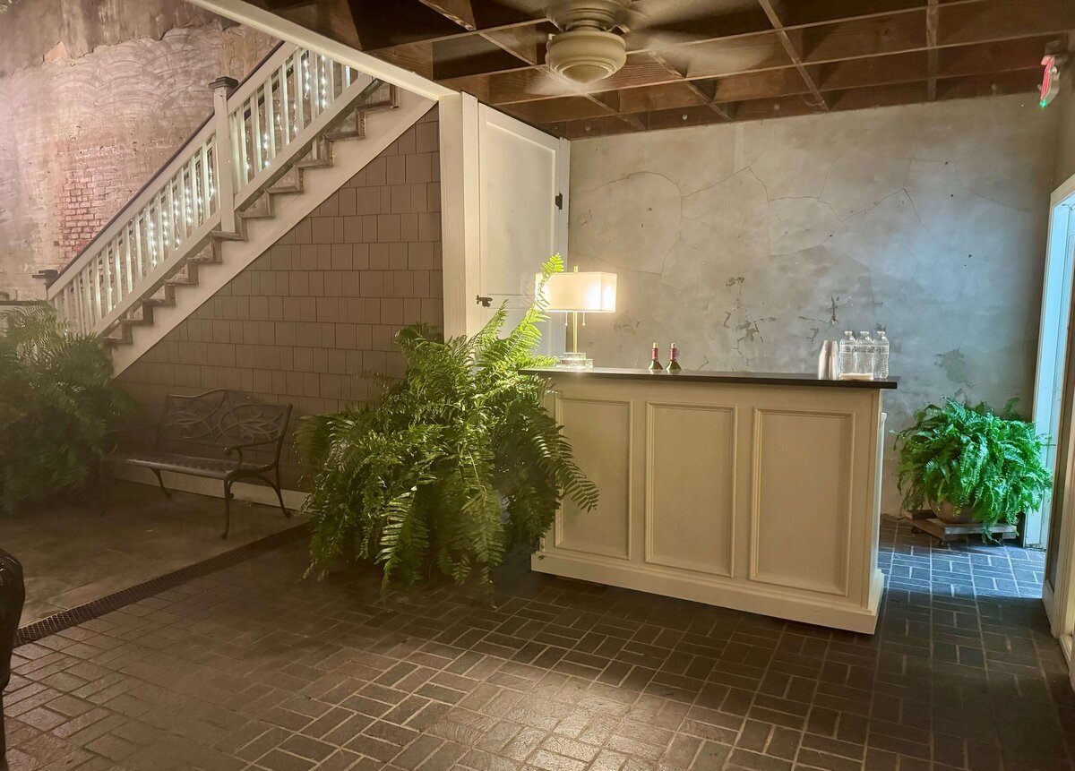 A new outdoor bar with a lamp, water pitcher, and glasses stands near a staircase and lush green ferns in a room with exposed brick and weathered walls. A bench is placed under the stairs.