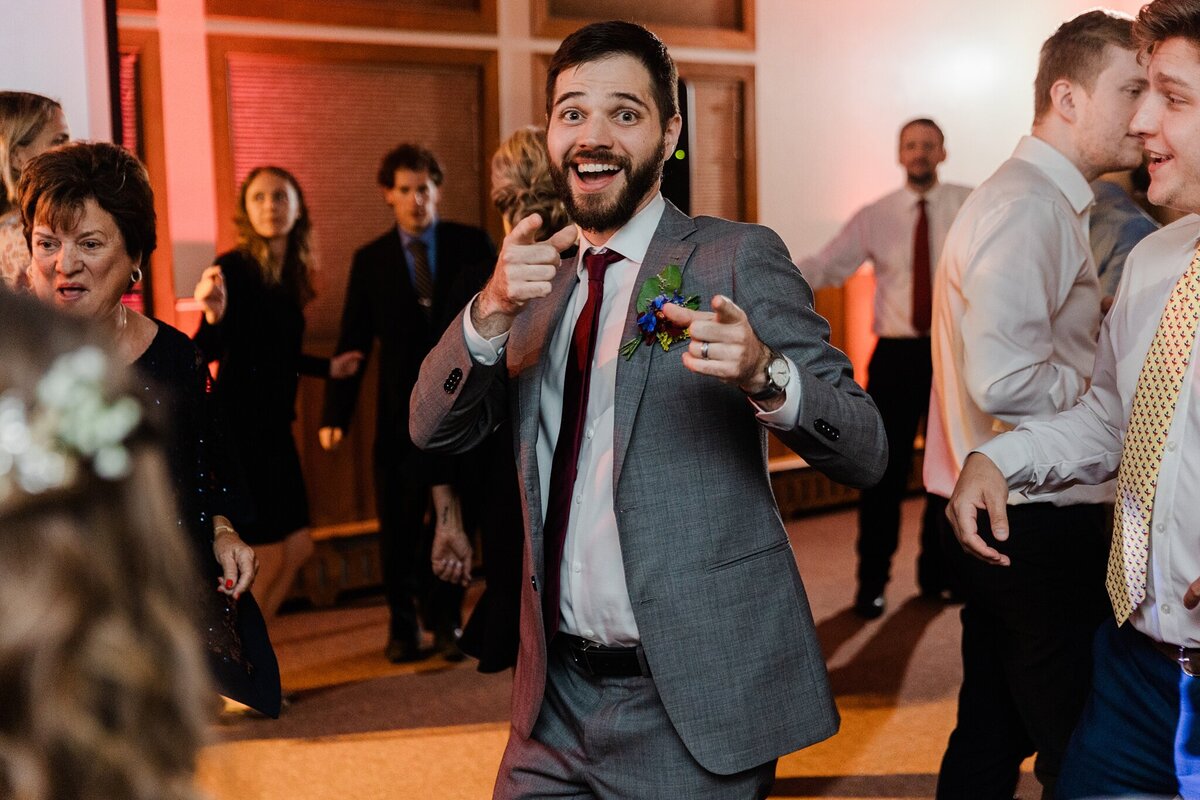 Photo of a groom pointing towards and looking playfully at the camera while dancing during his wedding reception at the YMCA of the Rockies in Estes Park, Colorado. The groom is wearing a gray suit with a boutonniere. Wedding guests can be seen dancing and celebrating all around him.
