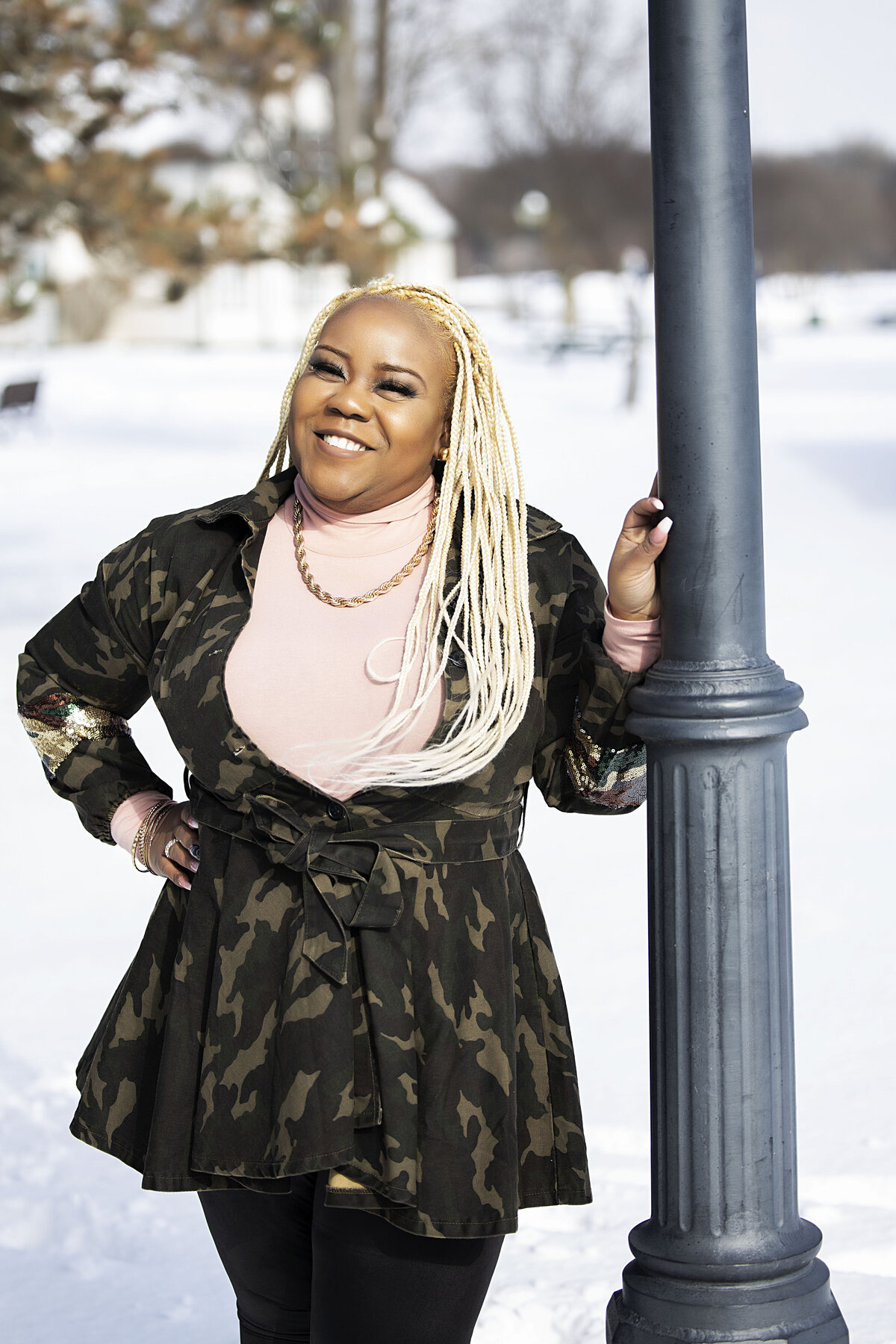 African American woman in camouflage jacket, pink turtleneck brown boots, and black jeans leaning on light pole