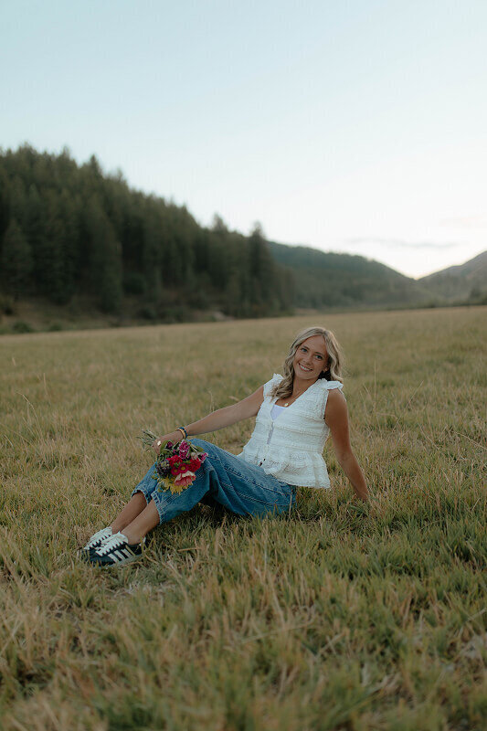Sitting on the ground in a grass field, a girl poses for her senior photos. Behind her is the Idaho Falls, Idaho landscape of hills and trees.