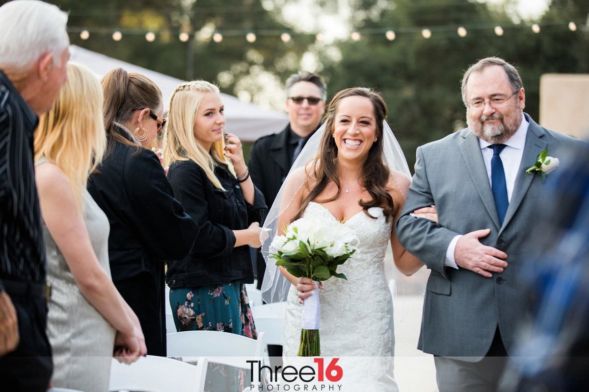 Bride is being escorted up the aisle by her father with her guests standing and she has a big grin on her face