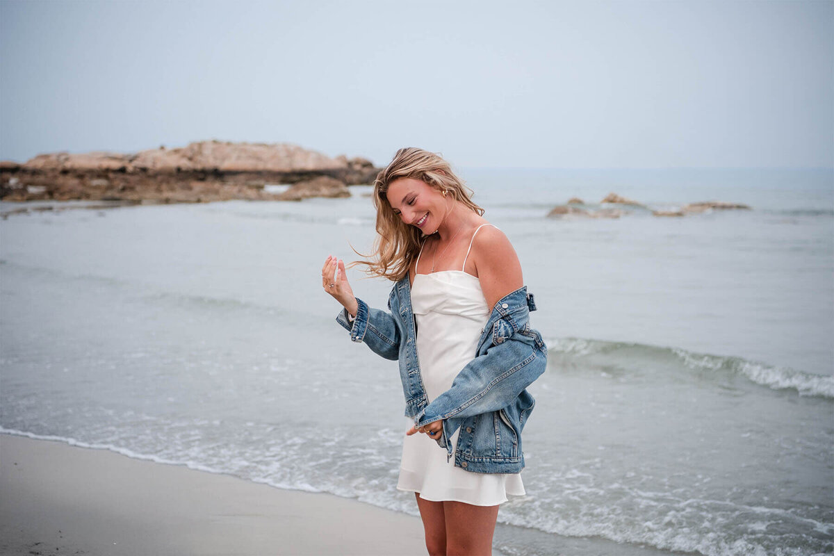 A senior stands on the North Shore beach of Wingaersheek in a dress and jean jacket as she laughs and the wind blows her hair.