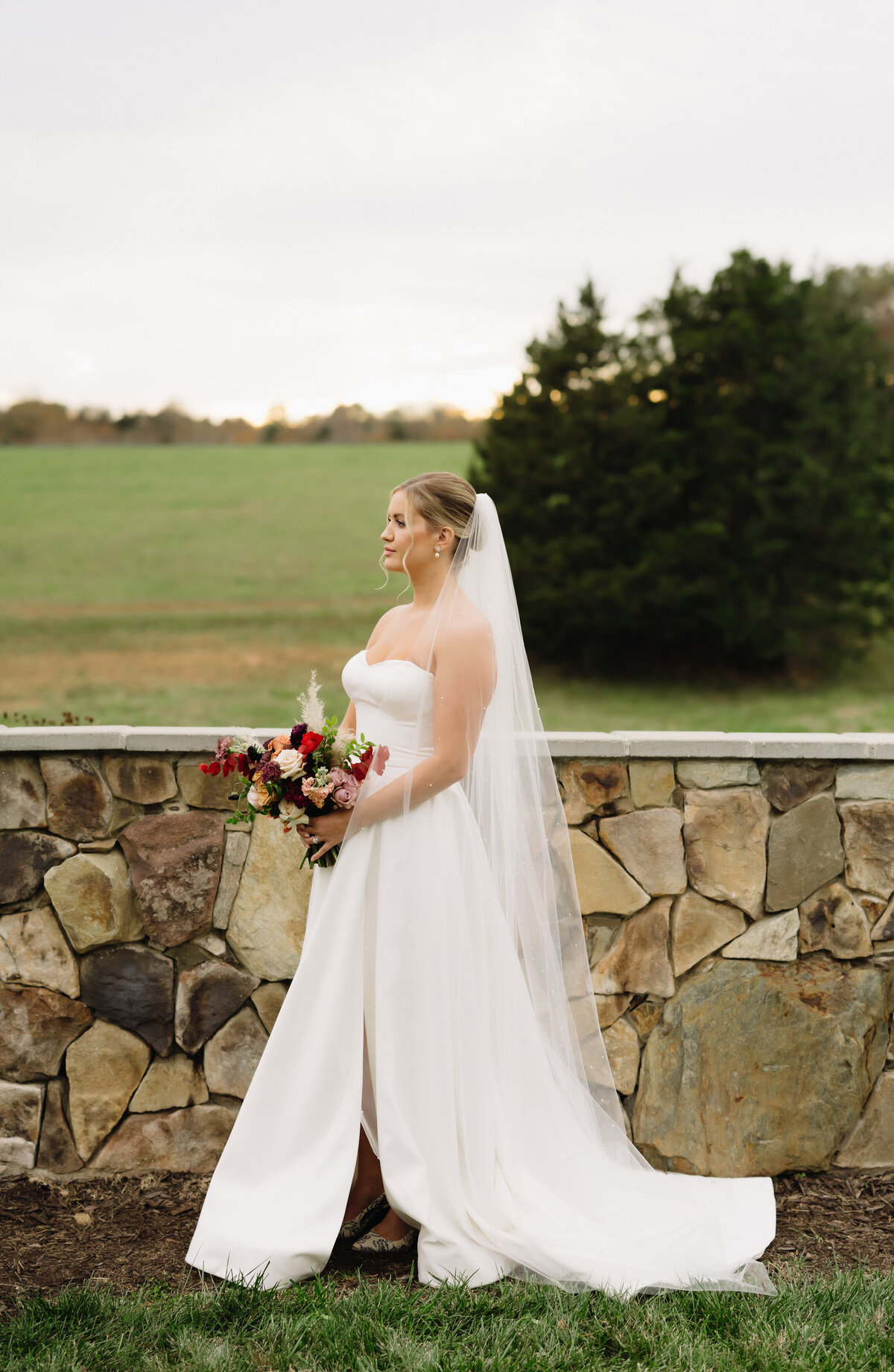outdoor wedding photo with bride with blonde hair in a bun with her veil flowing behind her as she holds her wedding bouquet with warm reds and pink flowers with a slit on the side of her gown