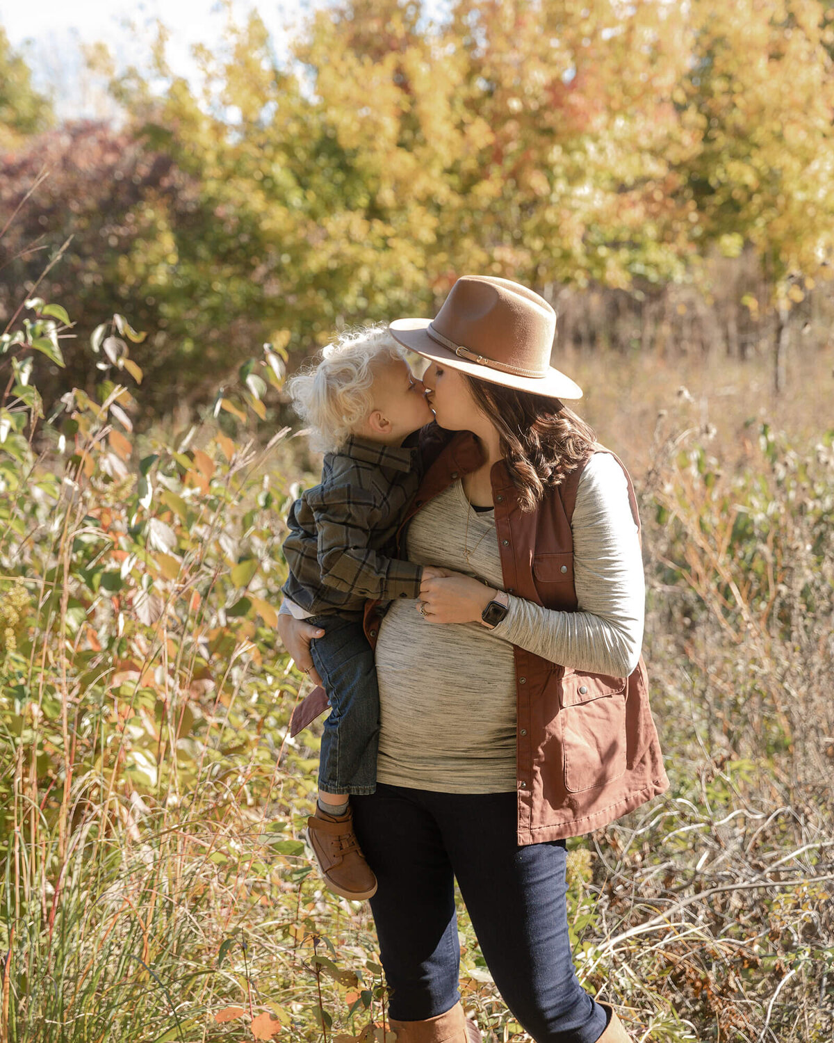 Pregnant Mom holding her toddler son and giving him a kiss in an open field.