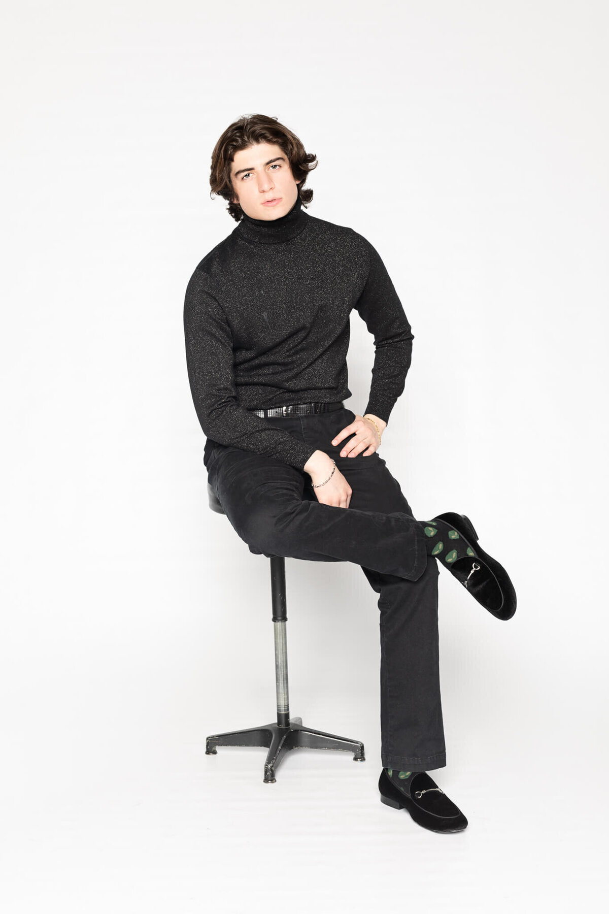 A person with medium-length brown hair is sitting on a stool against a plain white background. They are wearing a black turtleneck, black pants, and black shoes with green patterned socks. Their right leg is crossed over their left.