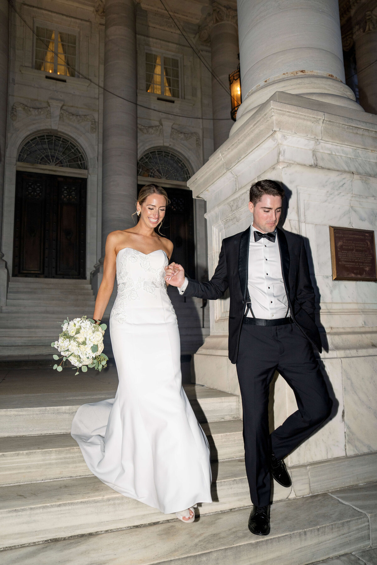A couple in formal attire descends marble steps. The woman, in a white wedding dress holding a bouquet, and the man, in a black tuxedo, are smiling under soft evening lighting. The background features grand architectural columns.