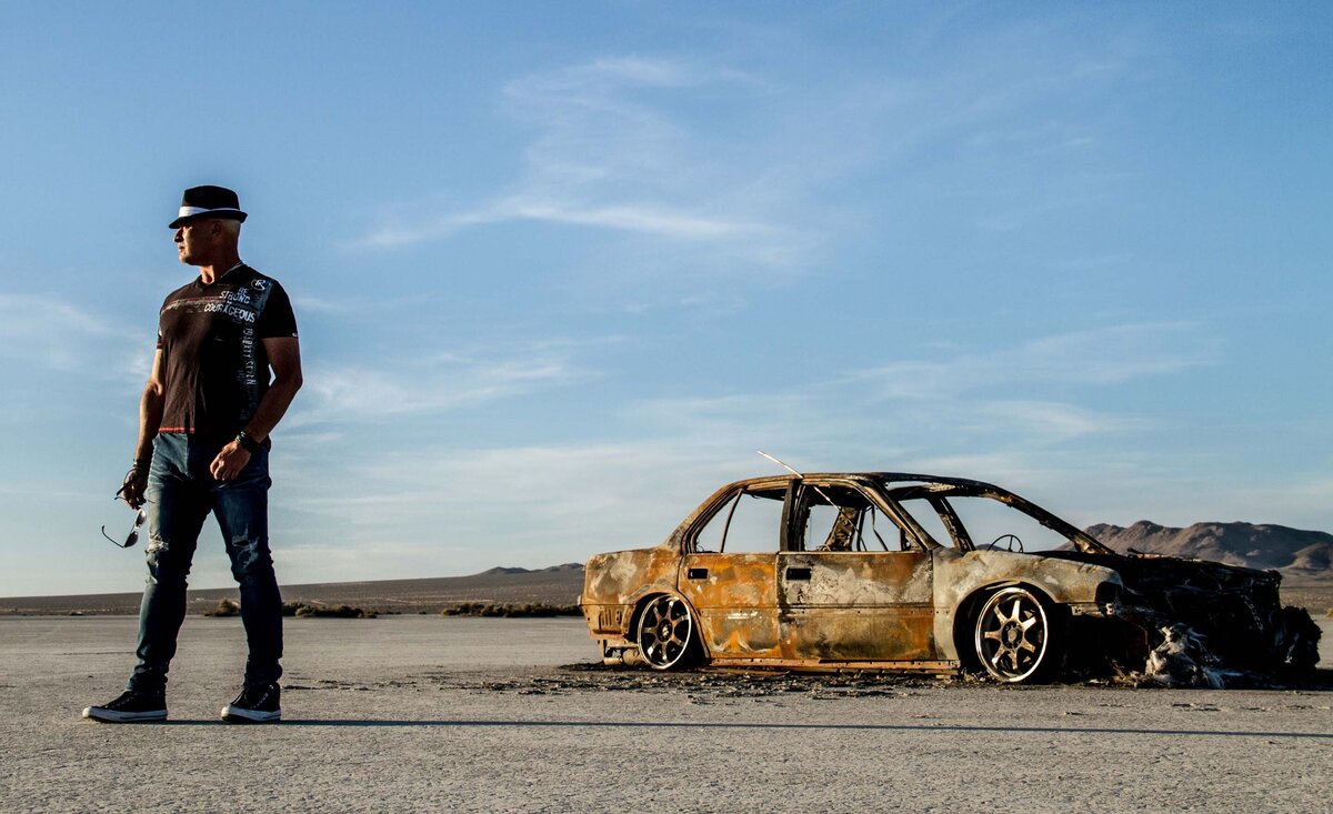 Male musician photo Adam Exler wearing black cloths black fedora standing beside burnt out car desert background