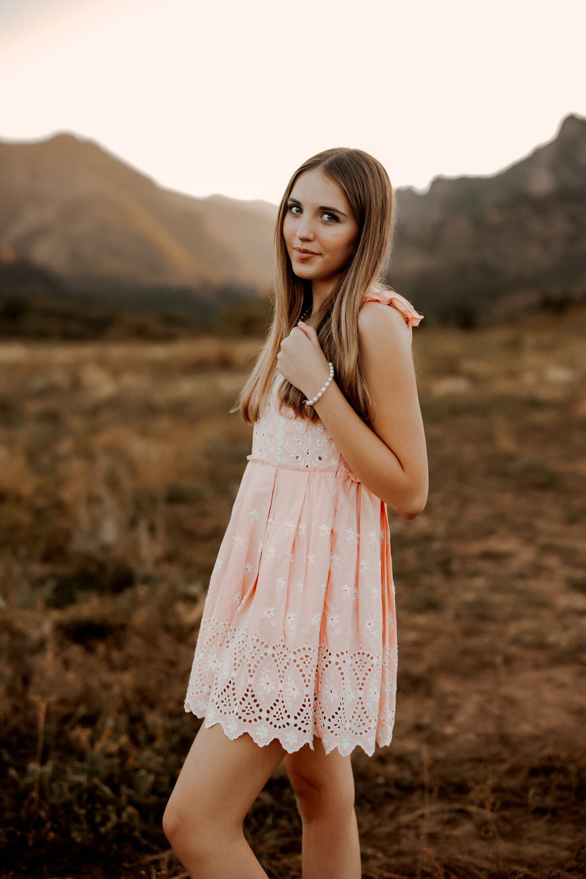 Senior girl in peach dress with long blond hair stands in open area with mountains behind her