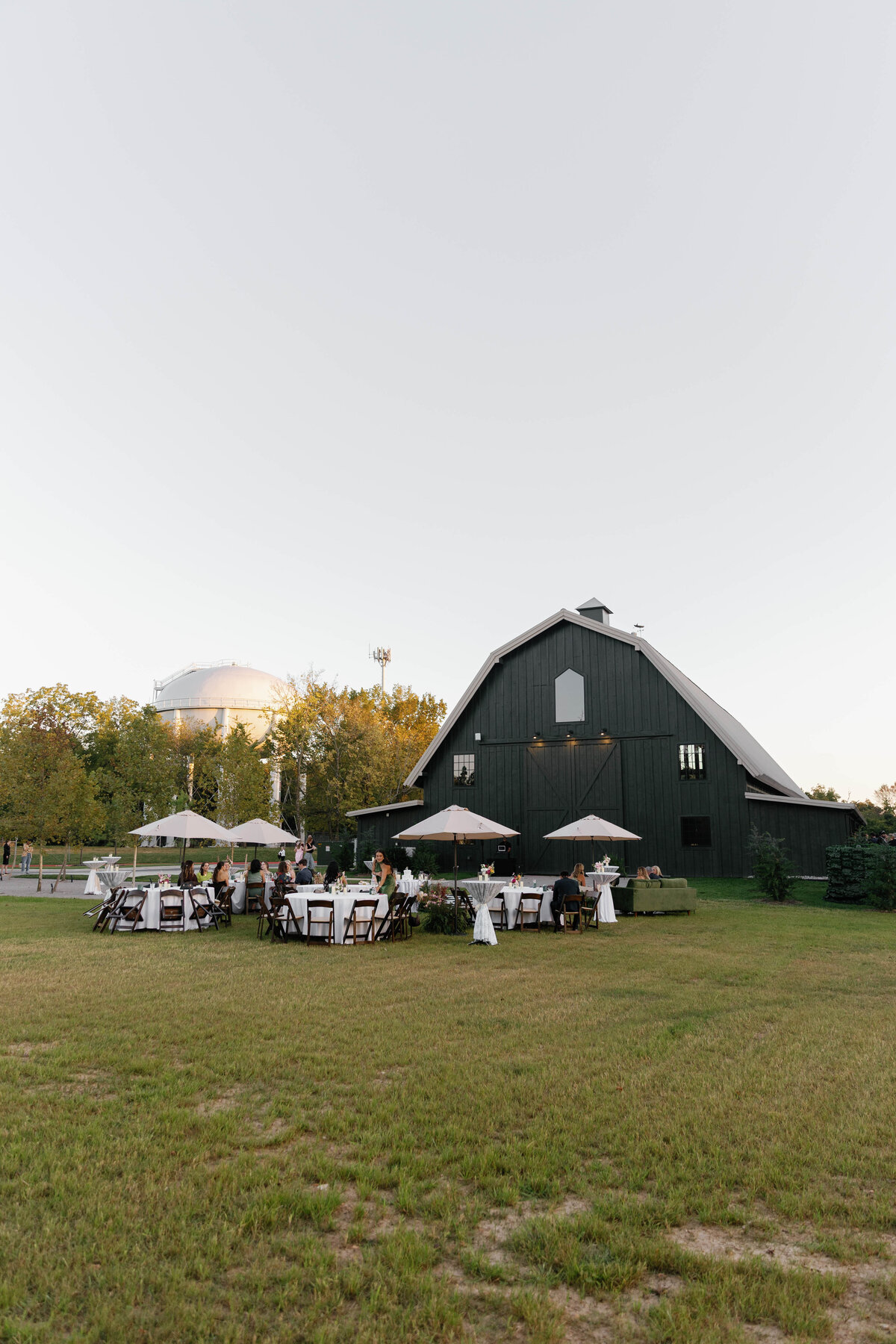 Open-air wedding reception beside a dark green barn at sunset, with round tables draped in white linens, floral centerpieces featuring blush and pastel blooms, and umbrellas for guest seating. Surrounded by golden light and trees, the setting showcases romantic outdoor wedding décor created with soft garden-style floral arrangements.