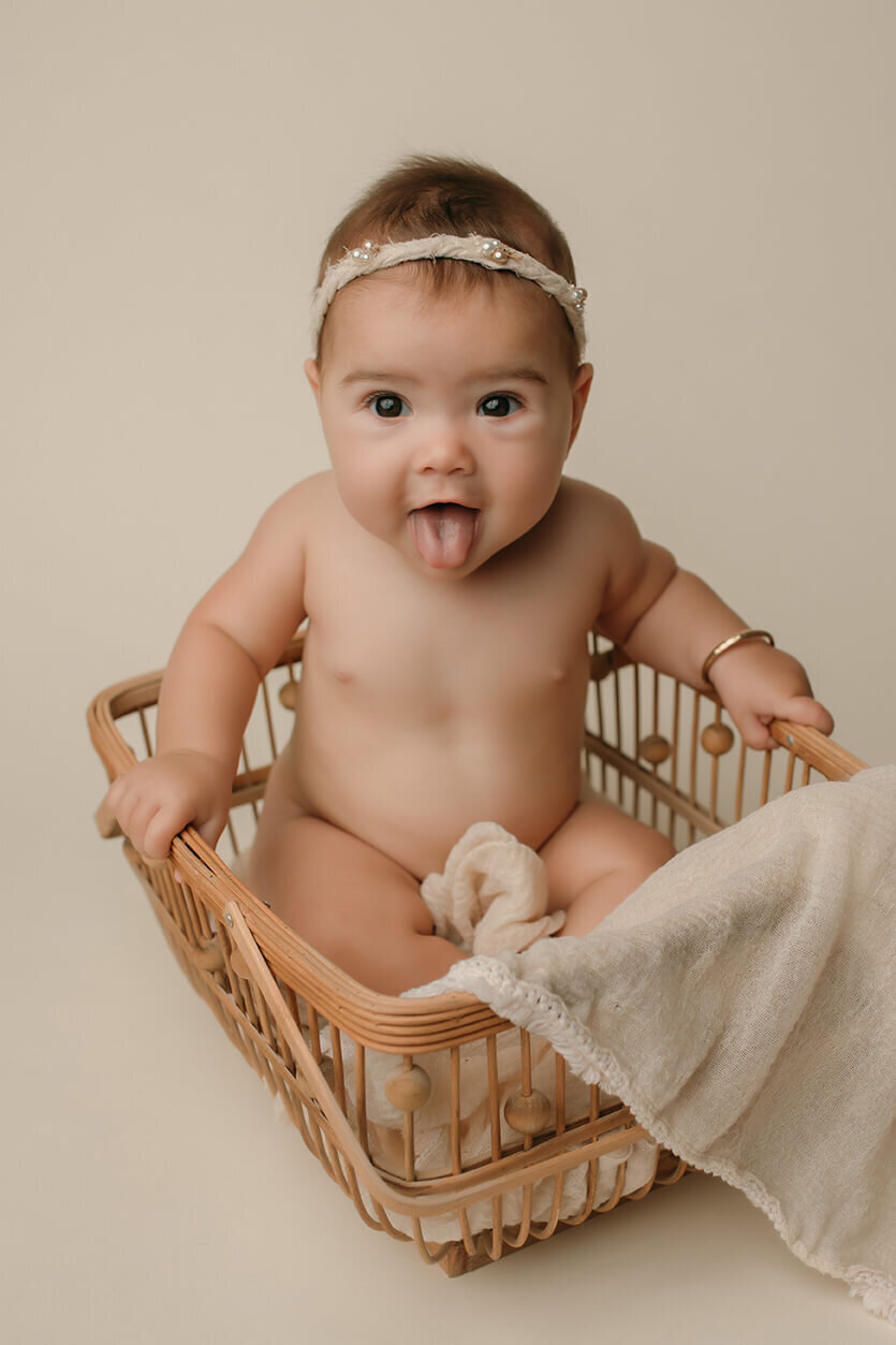 a six month old girl being silly in a wooden basket sticking her tongue out